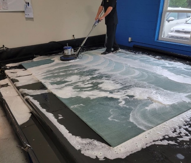Technician cleaning an area rug with a rotary floor machine at GM Carpet Care’s Toms River facility, using eco-friendly foam and specialized equipment for deep rug washing.